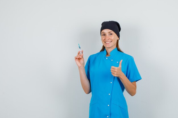 Doctora sosteniendo la jeringa con el pulgar hacia arriba en uniforme azul, sombrero negro y mirando alegre. vista frontal.
