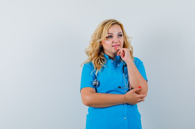 Doctora sosteniendo la mano en la barbilla mientras mira a un lado en uniforme azul y mira concentrada. espacio para texto