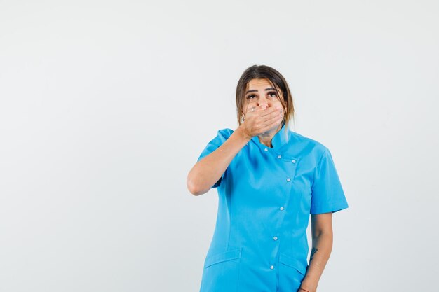 Doctora sosteniendo la mano en la boca en uniforme azul y mirando asustada