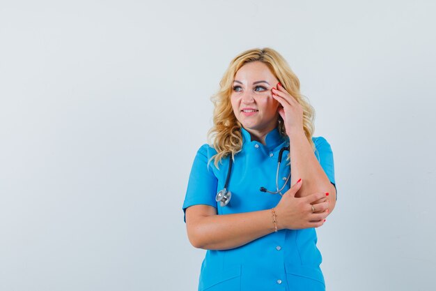 Doctora sosteniendo la mano en la sien mientras mira a un lado en uniforme azul y parece complacida. espacio para texto