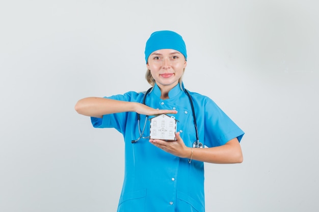 Doctora sosteniendo el modelo de la casa y sonriendo en uniforme azul