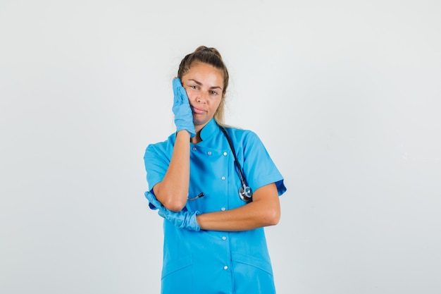 Doctora sosteniendo la palma en la mejilla en uniforme azul, guantes y mirando pensativo.