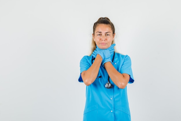 Doctora sosteniendo su garganta inflamada en uniforme azul, guantes y luciendo dolorosa