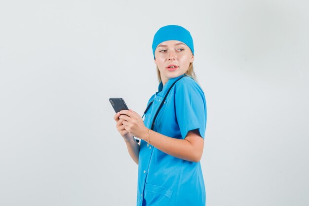 Doctora sosteniendo teléfono inteligente y mirando a un lado en uniforme azul y luciendo lindo