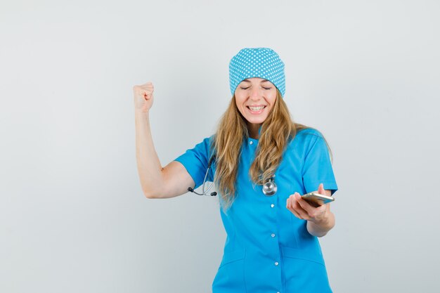 Doctora sosteniendo teléfono móvil con gesto de ganador en uniforme azul y mirando feliz.