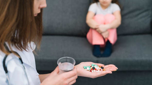 Doctora sosteniendo un vaso de agua y medicinas en la mano de pie delante de una niña enferma sentada en el sofá
