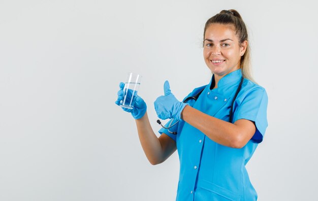 Doctora sosteniendo un vaso de agua con el pulgar hacia arriba en uniforme azul