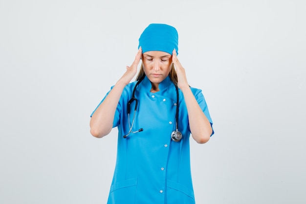 Doctora tocando las sienes con los dedos en uniforme azul y luciendo doloroso