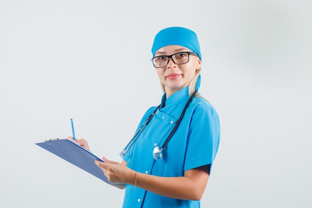 Doctora tomando notas en el portapapeles en uniforme azul, gafas y mirando alegre.