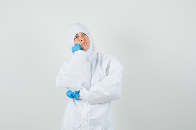 Doctora en traje de protección, guantes de pie en pose de pensamiento y mirando tranquilo