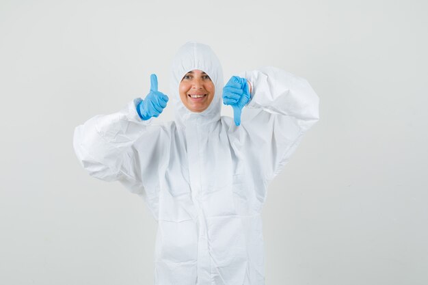 Doctora en traje protector, guantes mostrando los pulgares hacia arriba y hacia abajo y mirando contento