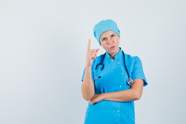 Doctora en uniforme azul advirtiendo con el dedo y mirando enojado, vista frontal.
