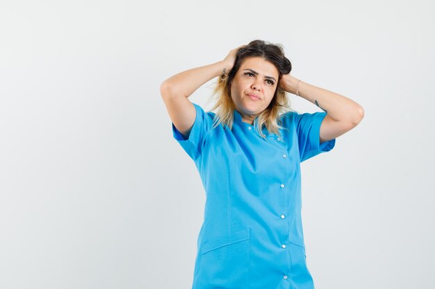 Doctora en uniforme azul agarrando la cabeza con las manos y mirando pensativo