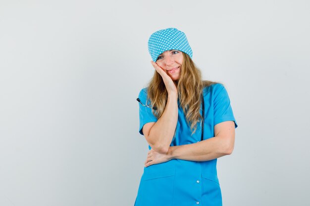 Doctora en uniforme azul apoyándose en la mejilla en la palma levantada y mirando soñadora