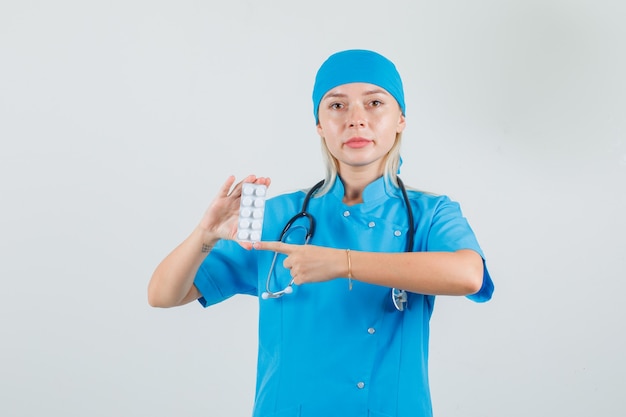 Doctora en uniforme azul apuntando al paquete de píldoras y mirando serio