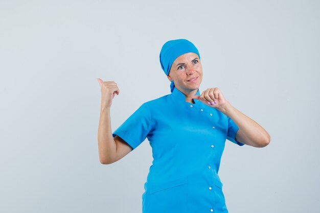 Doctora en uniforme azul apuntando hacia atrás con los pulgares y mirando esperanzado, vista frontal.