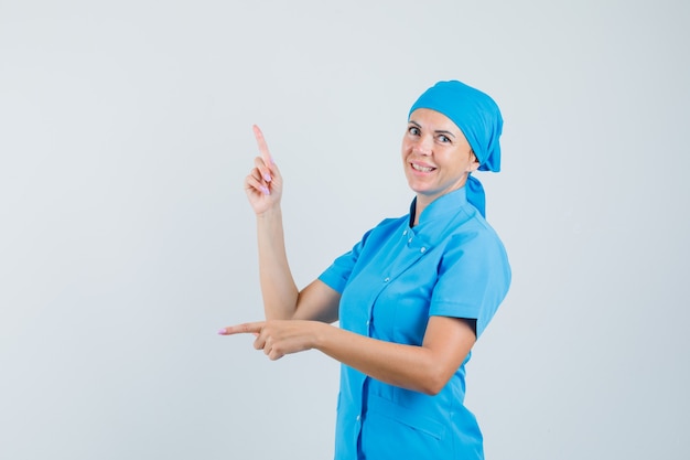 Doctora en uniforme azul apuntando con el dedo hacia arriba y hacia abajo y mirando vacilante, vista frontal.