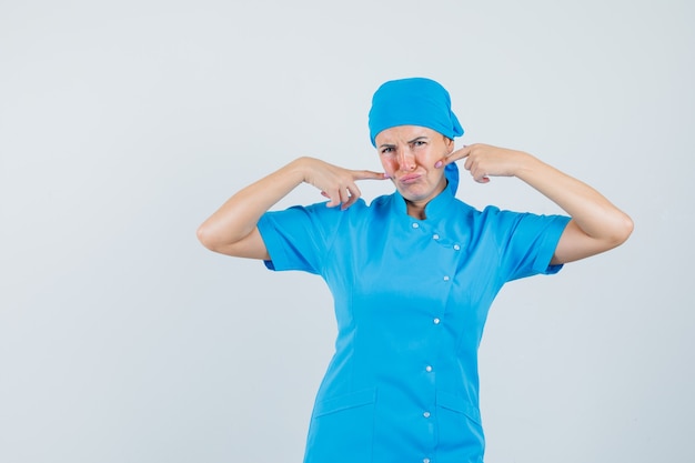 Doctora en uniforme azul apuntando con el dedo a las mejillas y mirando triste, vista frontal.