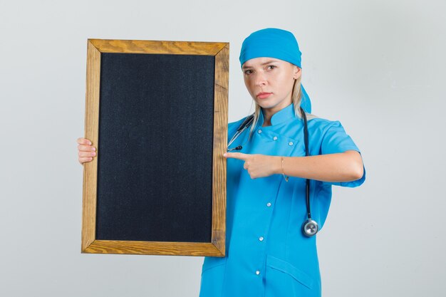 Doctora en uniforme azul apuntando con el dedo a la pizarra y mirando serio