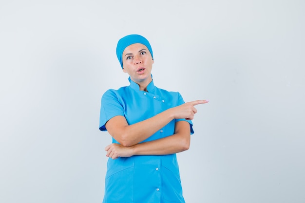 Doctora en uniforme azul apuntando hacia un lado y mirando sorprendido, vista frontal.