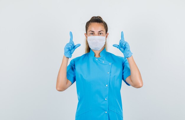 Doctora en uniforme azul, máscara, guantes apuntando hacia arriba con gesto de pistola y mirando confiado