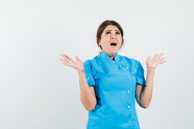 Doctora en uniforme azul mirando hacia arriba, levantando las manos, abriendo la boca y mirando indefenso