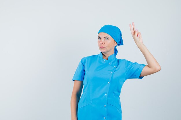 Doctora en uniforme azul mostrando gesto de victoria y mirando confiado, vista frontal.