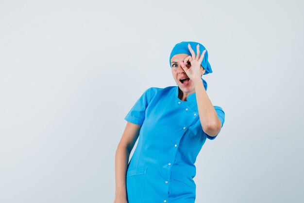 Doctora en uniforme azul mostrando signo ok en el ojo y mirando asombrado, vista frontal.