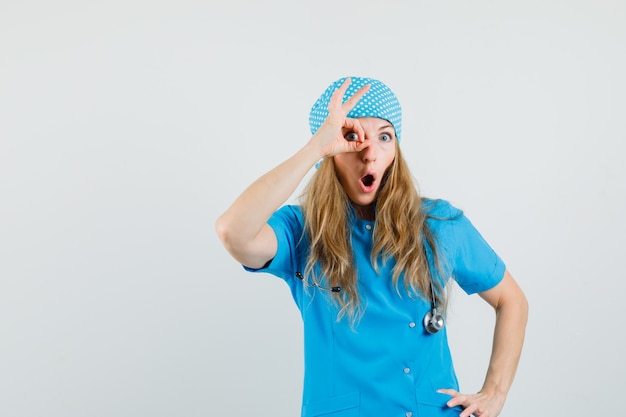 Doctora en uniforme azul mostrando signo ok en el ojo y mirando sorprendido