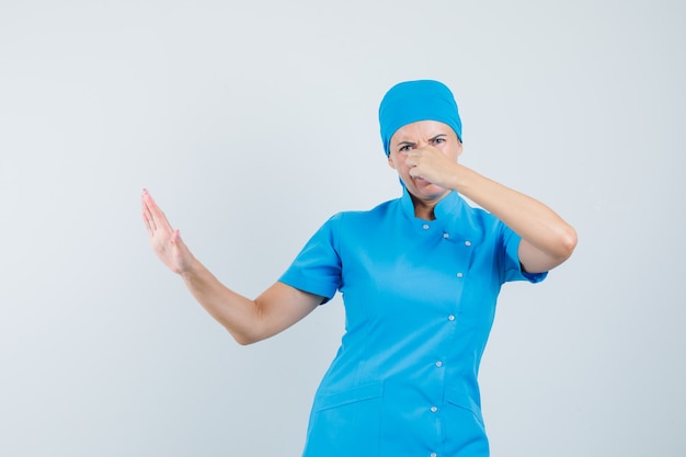 Doctora en uniforme azul pellizcando la nariz debido al mal olor y mirando disgustado, vista frontal.