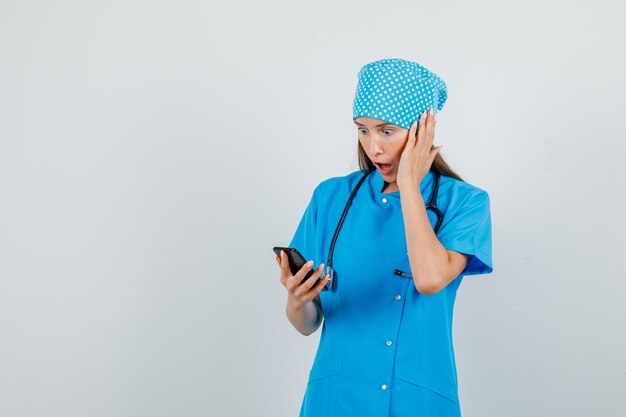 Doctora en uniforme azul con smartphone con la mano en la cara y mirando consternado