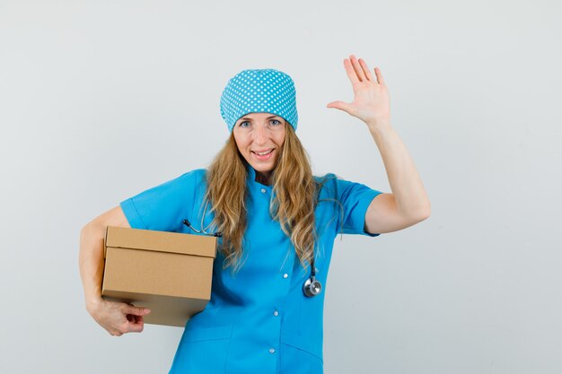 Doctora en uniforme azul sosteniendo una caja de cartón, agitando la mano y mirando alegre