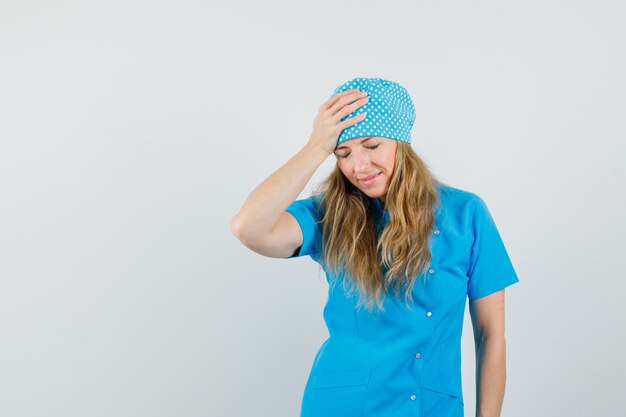 Doctora en uniforme azul sosteniendo la mano en la cabeza y mirando olvidadizo