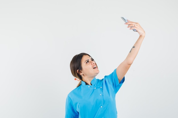 Doctora en uniforme azul tomando selfie en teléfono móvil y mirando alegre