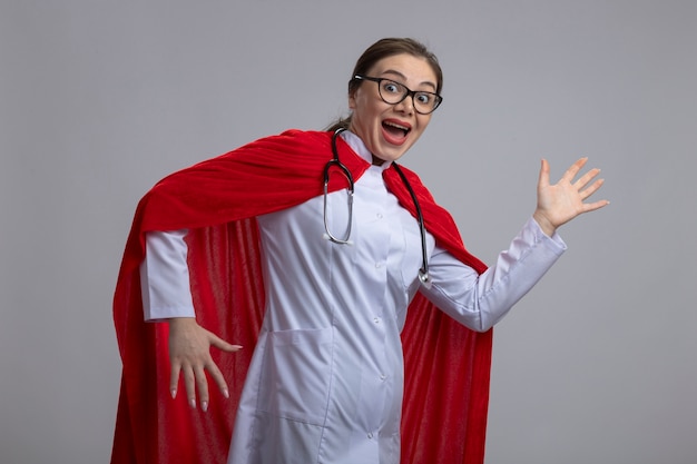 Doctora en uniforme blanco y capa roja de superhéroe que parece sorprendida y feliz lista para ayudar a pararse sobre la pared blanca