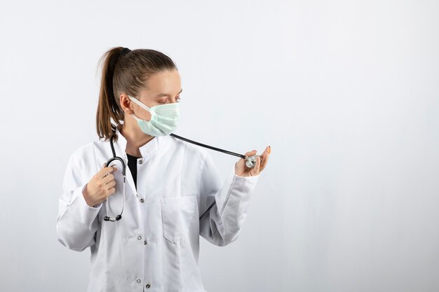 Doctora en uniforme blanco con una máscara médica y sosteniendo un estetoscopio