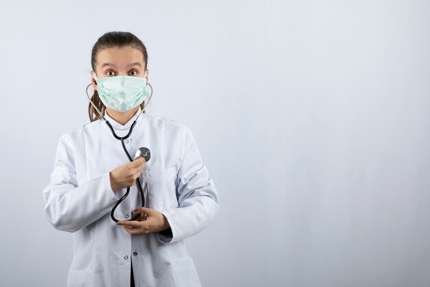 Doctora en uniforme blanco con una máscara médica y sosteniendo un estetoscopio