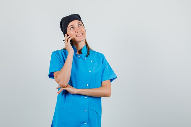 Doctora en uniforme mirando hacia arriba mientras habla por teléfono inteligente y se ve alegre