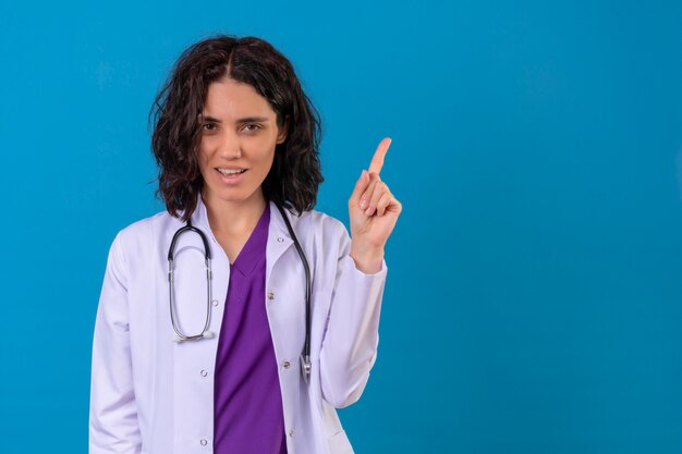 Doctora vistiendo bata blanca con estetoscopio apuntando hacia arriba con el dedo sonriendo confiado concentrado en la tarea de pie en azul aislado con espacio de copia