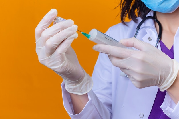 Doctora vistiendo bata blanca con estetoscopio en máscara protectora médica con jeringa y frasco de vacuna de frasco de medicina en naranja aislado