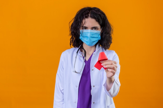 Foto gratuita doctora vistiendo bata blanca con estetoscopio en máscara protectora médica sosteniendo una cinta roja, un símbolo de la lucha contra el sida con cara seria de pie