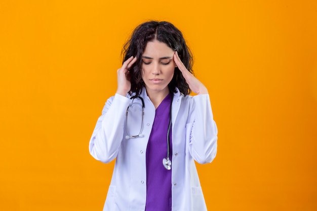 Doctora vistiendo bata blanca con estetoscopio mirando mal tocar templos con fuerte dolor de cabeza de pie en naranja aislada
