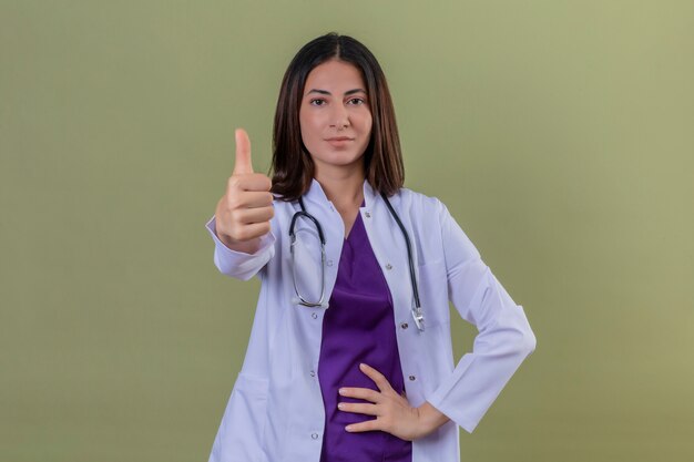 Doctora vistiendo bata blanca y con estetoscopio mirando seguro mostrando los pulgares para arriba en verde aislado