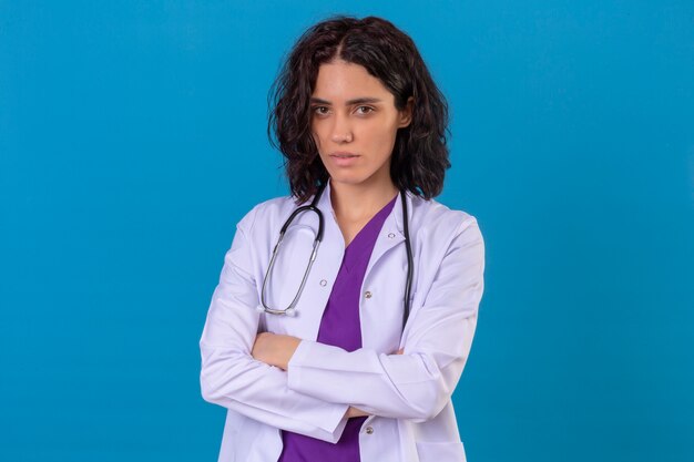 Doctora vistiendo bata blanca con estetoscopio de pie con los brazos cruzados con el ceño fruncido en azul aislado