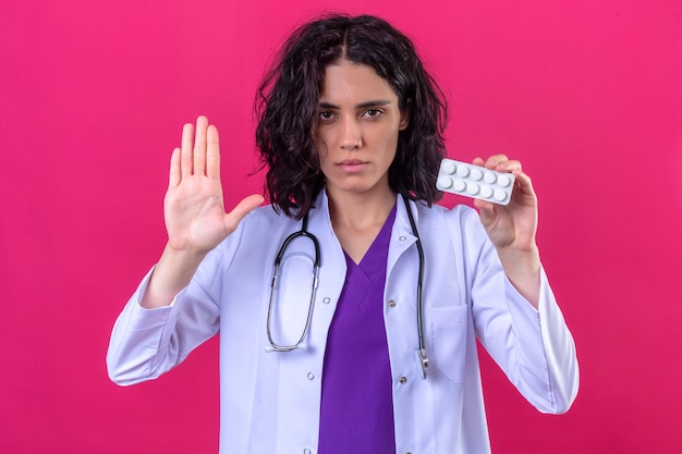 Foto gratuita doctora vistiendo bata blanca con estetoscopio de pie con la mano abierta haciendo gesto de parada y sosteniendo el blister de pastillas en la otra mano en rosa aislado