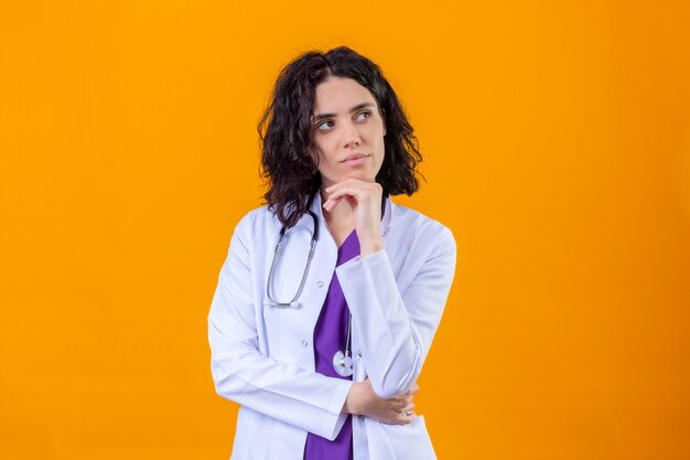 Doctora vistiendo bata blanca con estetoscopio de pie con la mano en la barbilla mirando a un lado pensando mirada soñadora de pie en naranja aislada