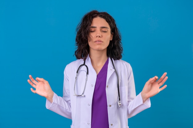 Doctora vistiendo bata blanca con estetoscopio relajarse y sonreír con los ojos cerrados haciendo gestos de meditación con los dedos en azul aislado