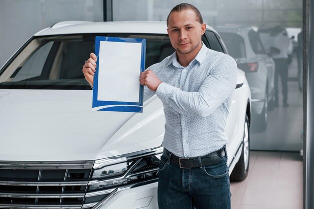 Documento Importante. Gerente se encuentra en frente del coche blanco moderno con papel en las manos