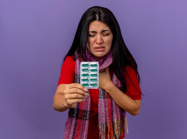 Doliente joven caucásica enferma con bufanda mostrando paquete de cápsulas poniendo la mano en el pecho con los ojos cerrados aislado en la pared púrpura con espacio de copia