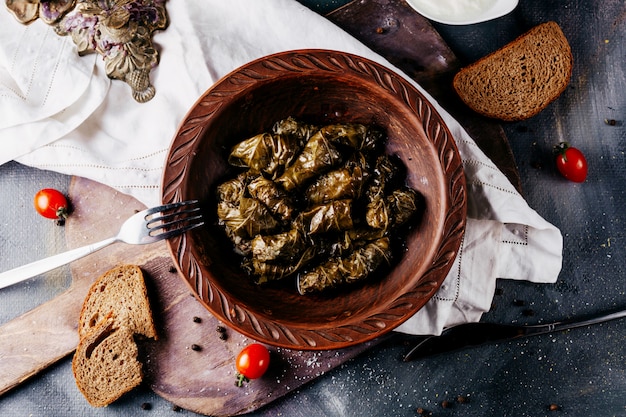 Dolma verde una vista superior dentro de la placa marrón alrededor de tomates cherry rojos y rebanadas de pan en la superficie oscura